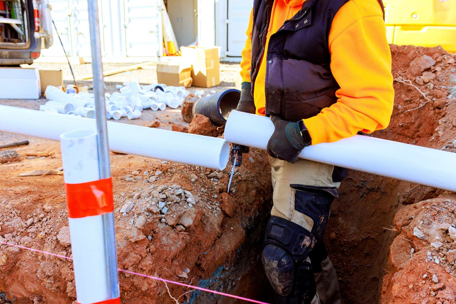 A construction worker in safety gear is cutting a white PVC pipe at a construction site, with pipes and equipment visible in the background.