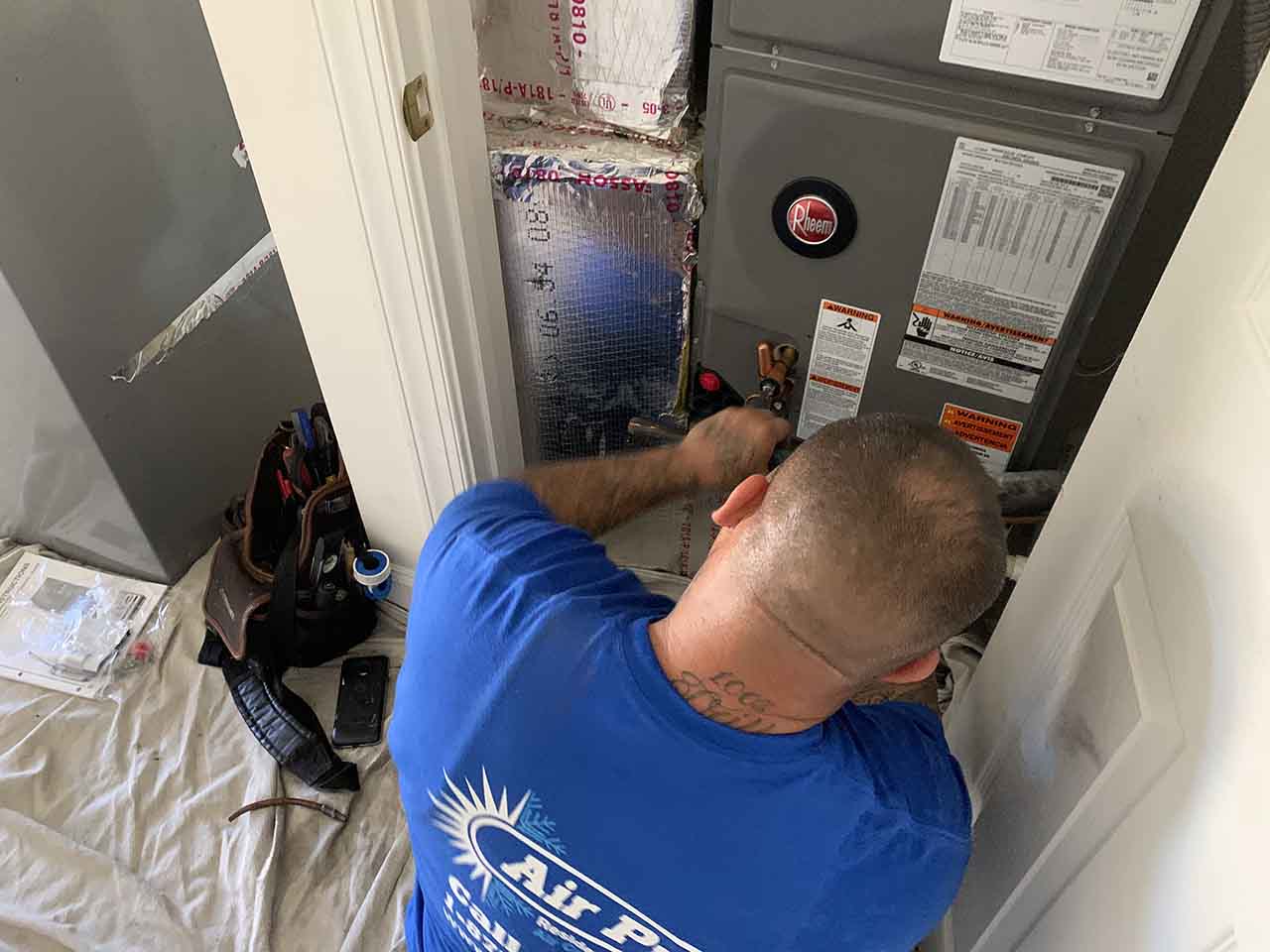 A technician in a blue shirt inspects a central AC unit inside a small utility closet, with tools and equipment on the floor nearby, troubleshooting issues like intermittent cold air.