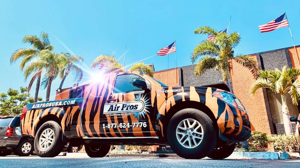A tiger-striped Air Pros service truck, specializing in AC repair and installs, is parked in front of a building with palm trees and three American flags.