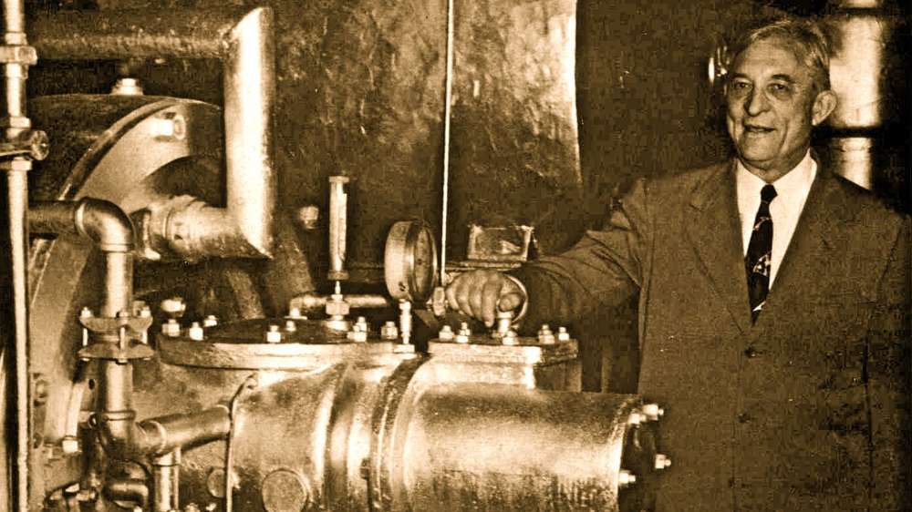 A man in a suit stands beside industrial machinery, smiling, with one hand resting on a large valve—highlighting the rich history of air conditioning and its impact on modern industry.