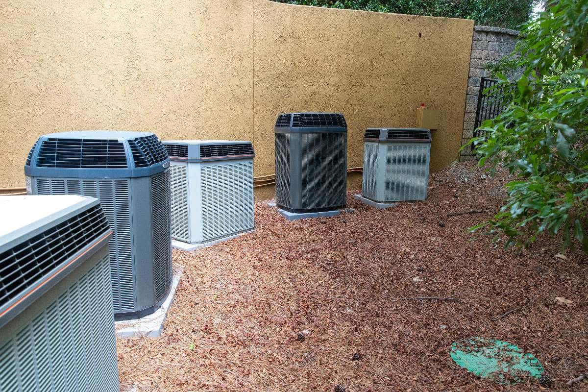Five outdoor central AC units are installed on the ground along a yellow stucco wall, surrounded by mulch and greenery, highlighting the importance of proper placement to maximize air conditioner lifespan.