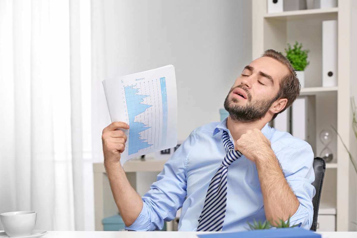 A man in an office uses a report with blue graphs to fan himself, loosening his tie and looking overheated—clearly wishing for better air balancing.