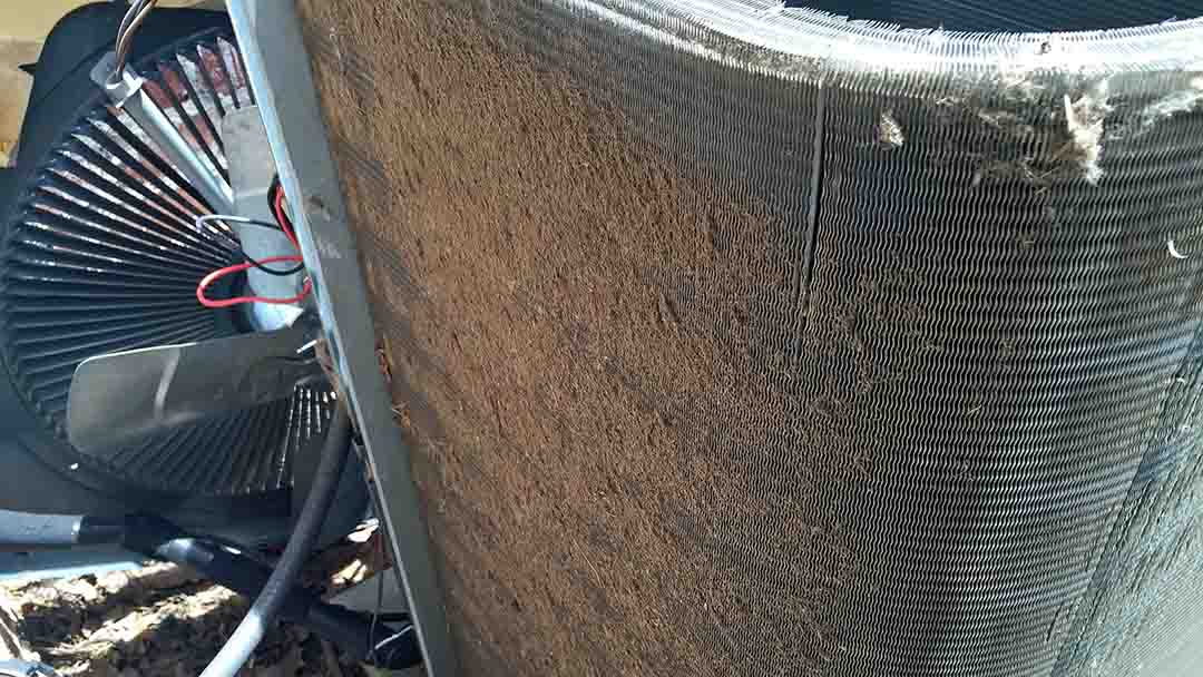 Close-up of a central AC unit outdoors with a heavily clogged condenser coil, showing dirt and debris buildup on the metal fins.