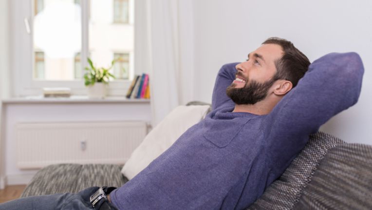 A man with a beard relaxes on a sofa with his hands behind his head, smiling and enjoying summer comfort in a bright, modern living room.