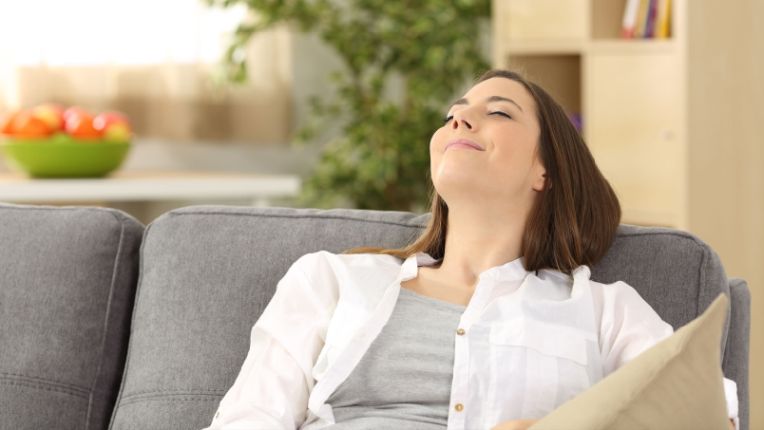 A woman sits on a gray sofa with her eyes closed and a slight smile, appearing relaxed—enjoying cool comfort and thinking about summer cooling tips. A bowl of fruit and plants are visible in the background.