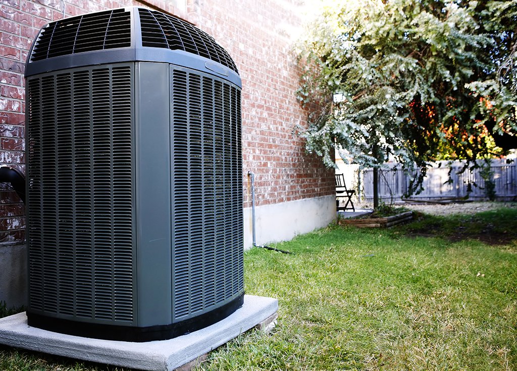 A large Central AC unit sits on a concrete pad beside a brick house, with green grass and a wooden fence visible in the background.