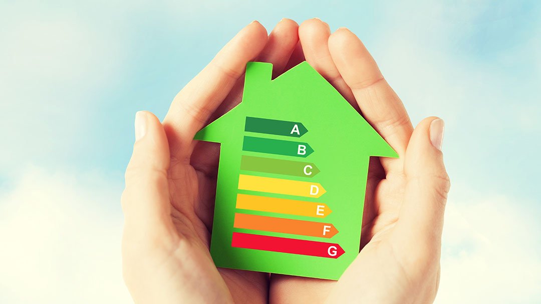Hands hold a green house-shaped cutout displaying an Energy Efficiency Rating (EER) chart from A (green) to G (red) against a blue sky background.