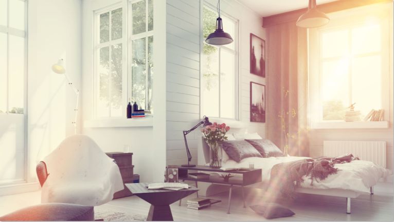 Bright bedroom with large windows, modern furniture, and a cozy bed with pillows. Sunlight streams in, while decorative plants and books line the shelves—perfect for warmer climates when looking to save money on AC repair.