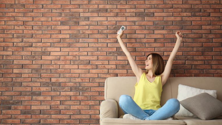 A woman in a yellow top sits cross-legged on a beige sofa, raising her arms and holding a remote, in front of a brick wall—enjoying energy efficiency after eliminating air leaks.