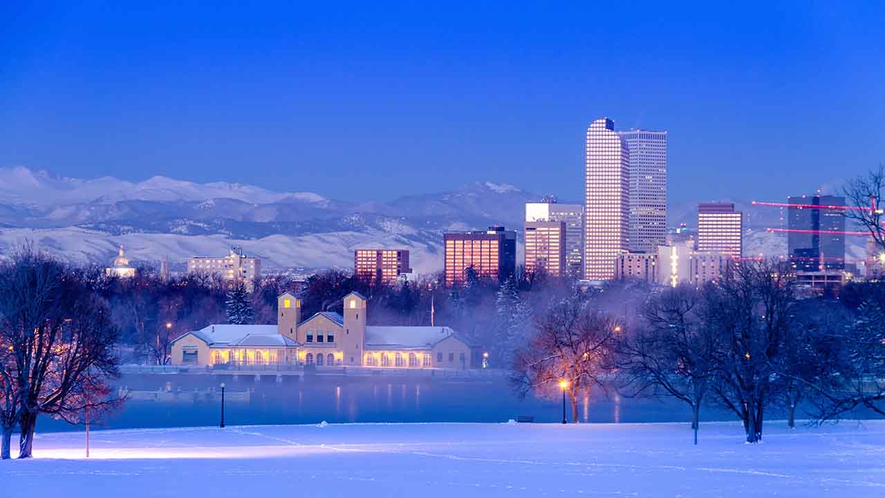 Snow-covered park with a large building by a lake in the foreground; city skyline and mountains visible under a clear twilight sky in the background, typical of the winter months—remember to protect central AC during this season.