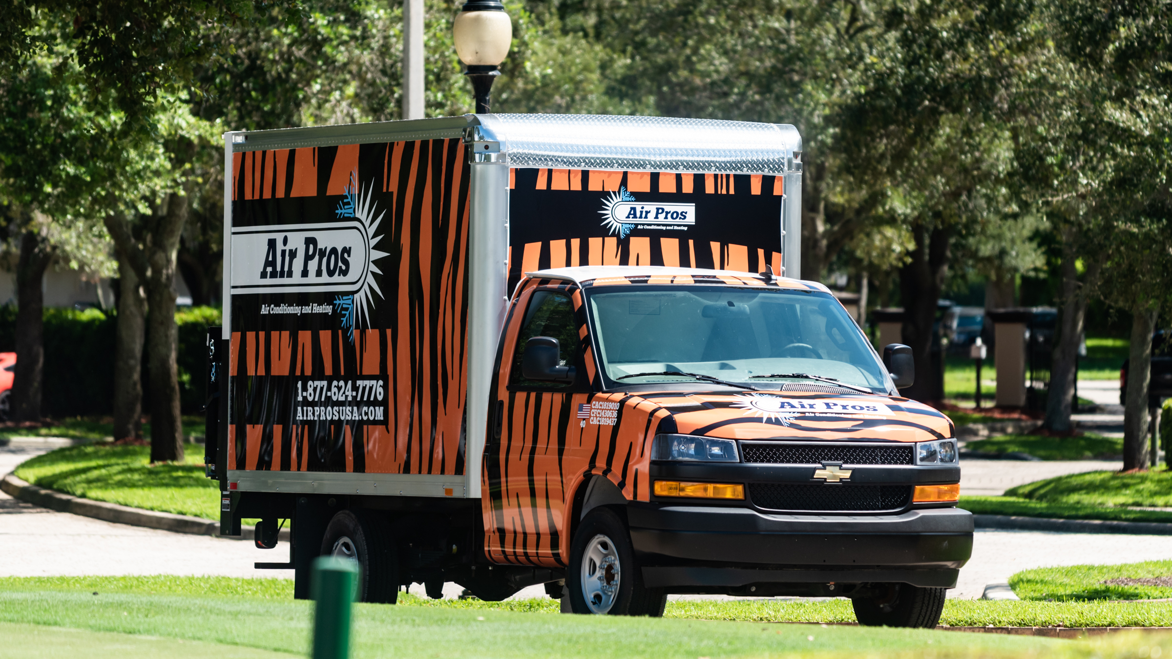 A box truck with tiger stripe graphics and "Air Pros USA" branding, part of a donation to the Boys & Girls Clubs of Broward County, is parked on a suburban street with trees and grass in the background.