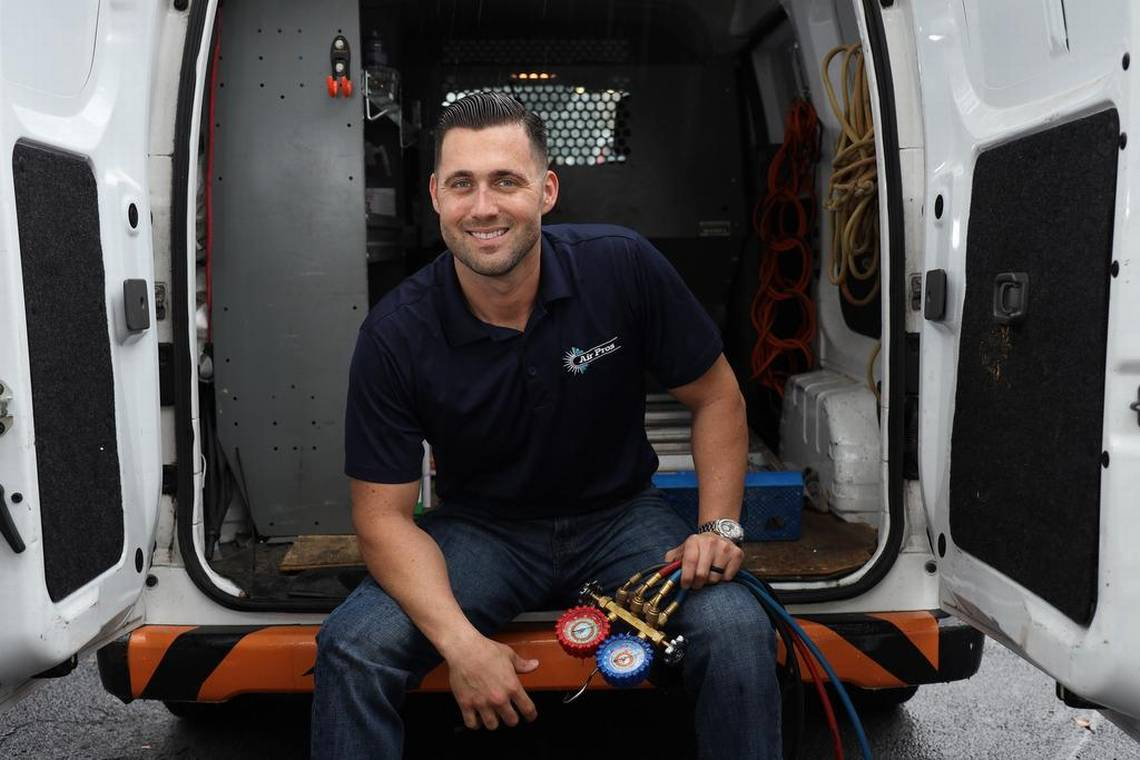 A man in a navy polo shirt sits in the open back of a work van, holding HVAC manifold gauges and smiling at the camera—representing Air Pros as they deliver top-notch service across Florida.