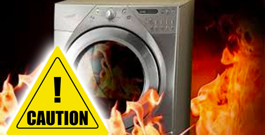 A front-loading washing machine is surrounded by flames, with a large yellow caution sign in the foreground—highlighting fire safety concerns and the risk of clothes dryer fires during the winter months.