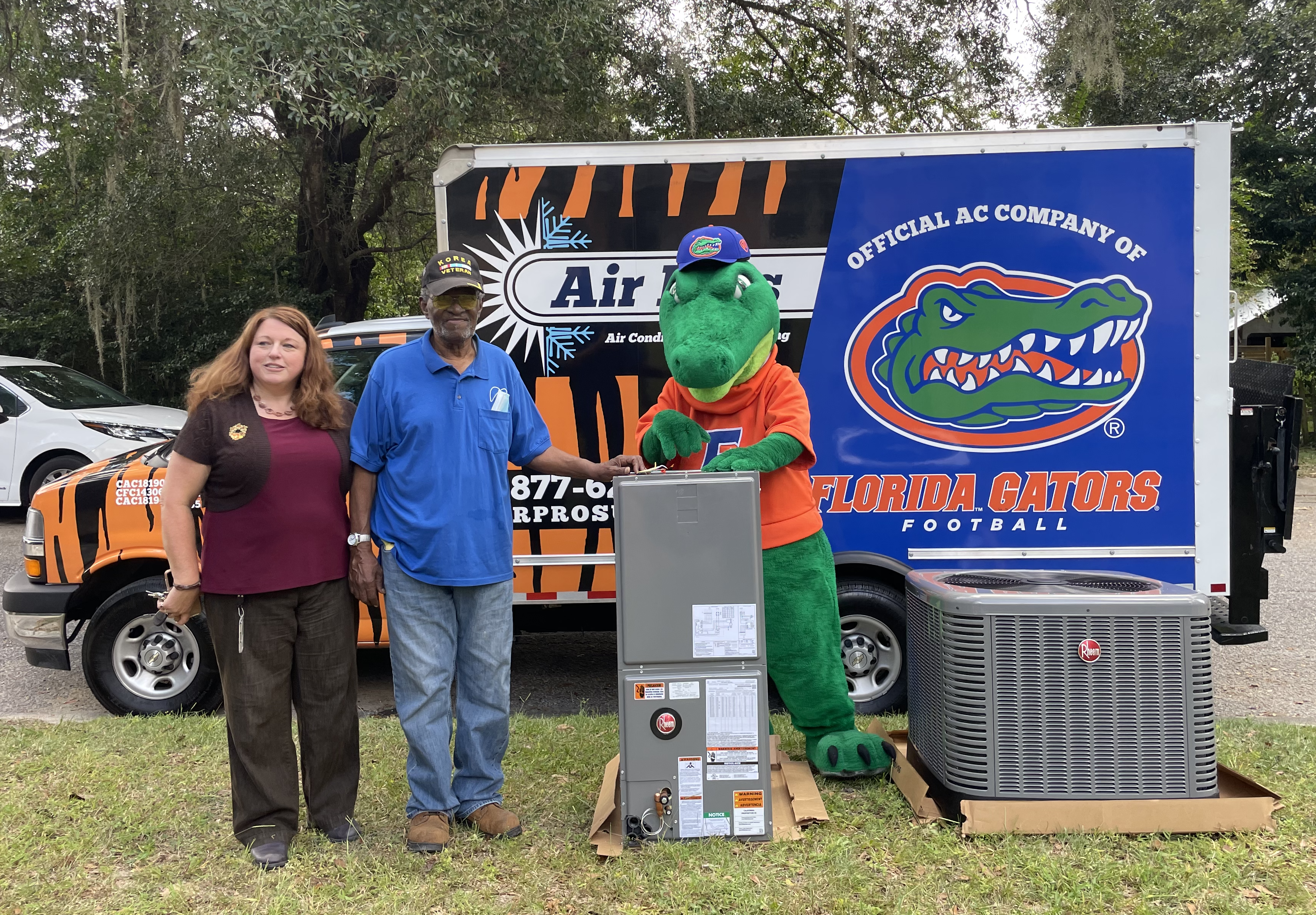 Two people and a person in an alligator mascot costume pose in front of an HVAC unit and a van with Florida Gators Football and Air Pros USA branding.