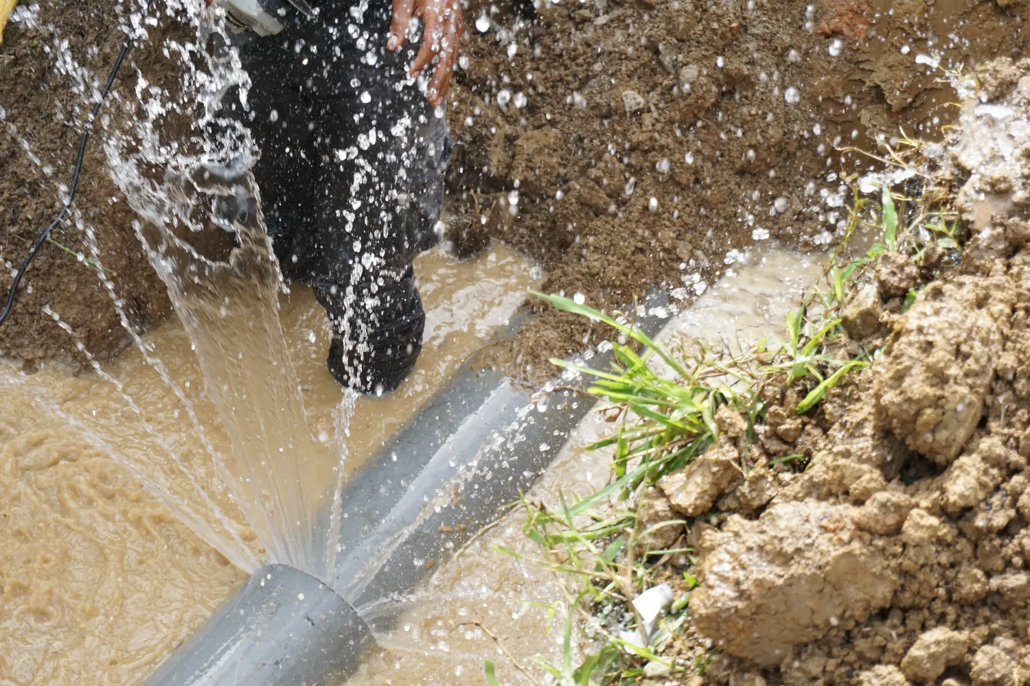 Water sprays from a burst pipe in a muddy trench as a worker attempts to repair the leak.