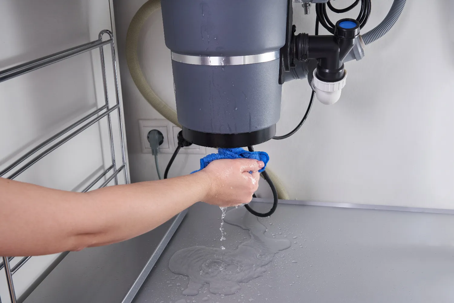 A person uses a cloth to catch leaking water from a pipe under a kitchen sink near the garbage disposal, with water spilled on the surface below.