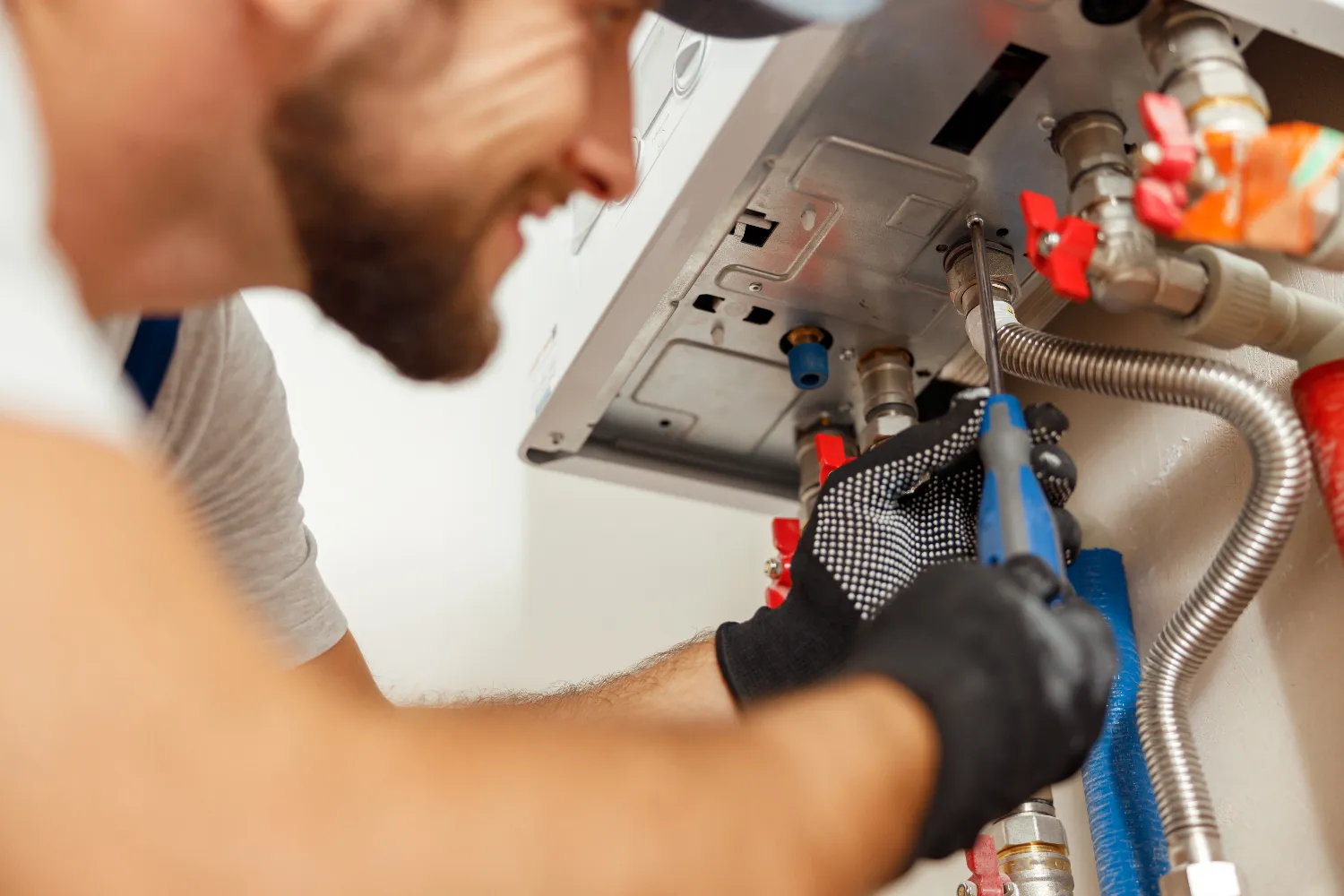 A plumber wearing gloves uses a wrench to adjust pipes connected to a heating or water system, specializing in Water Heater Installation and Repair.