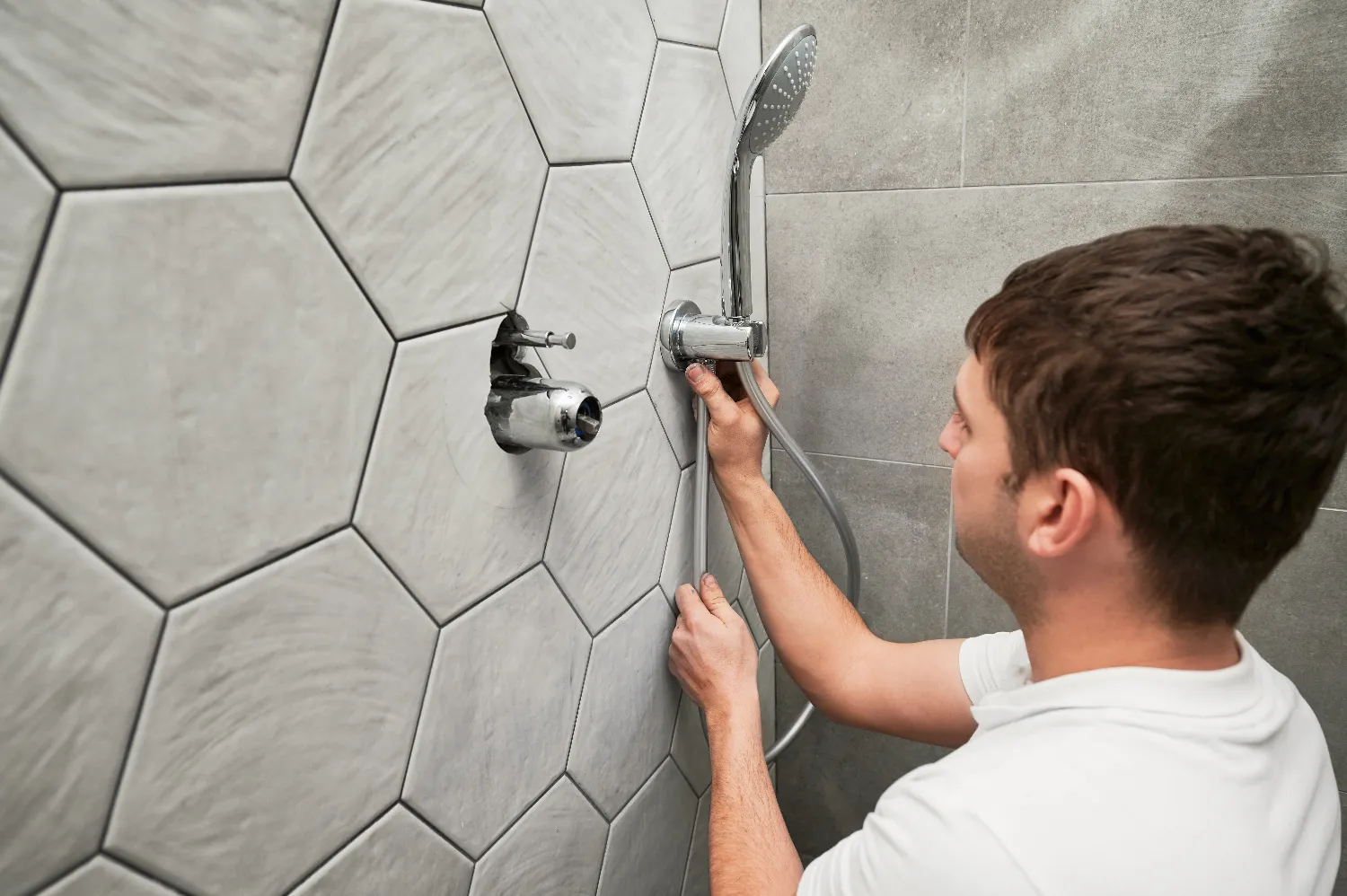 A person handles bathroom plumbing while installing or repairing a shower faucet and handheld showerhead on a bathroom wall with large hexagonal tiles.