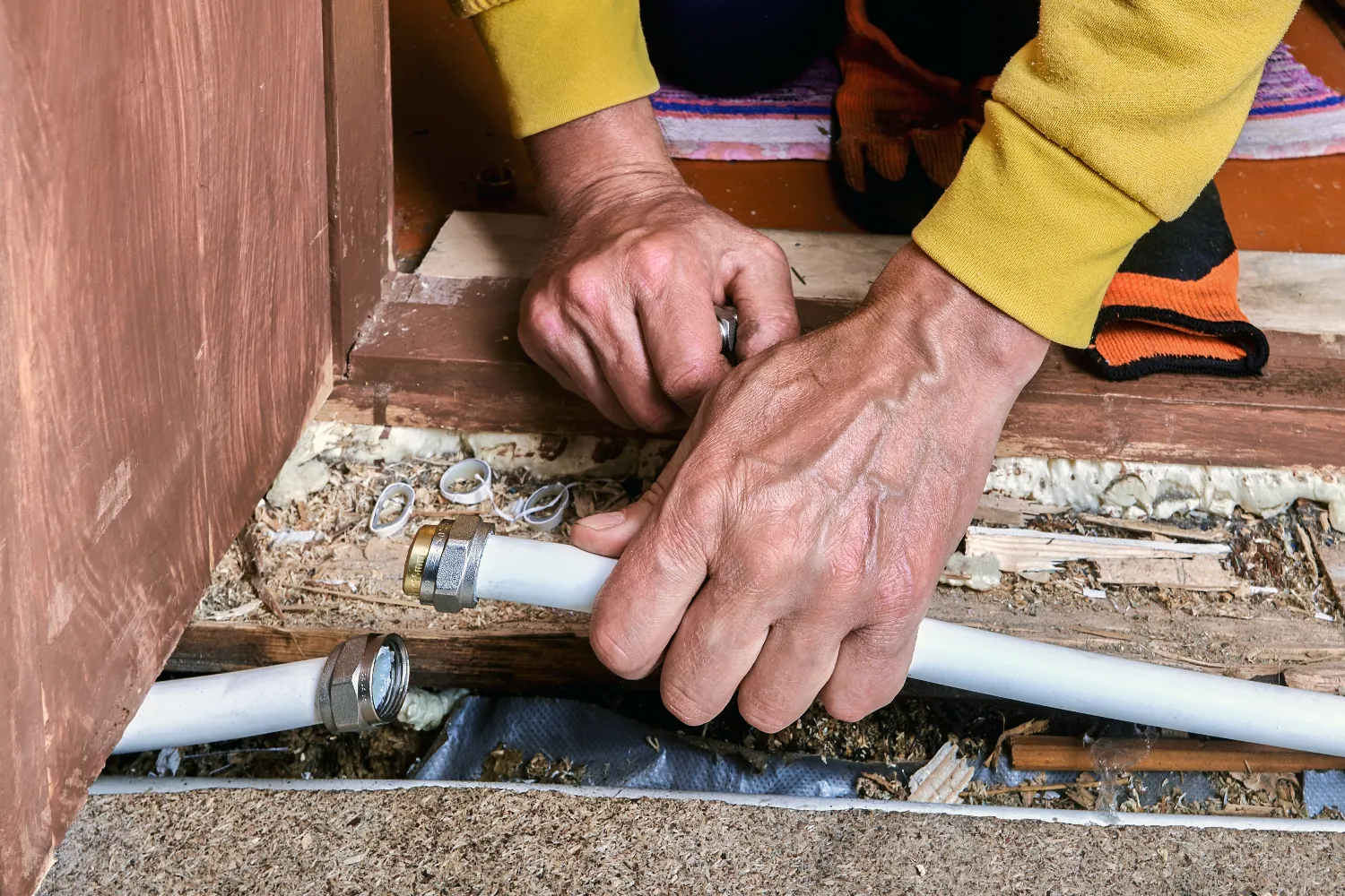 Person's hands connecting plastic plumbing pipes near a doorway with debris and tools visible around the work area.