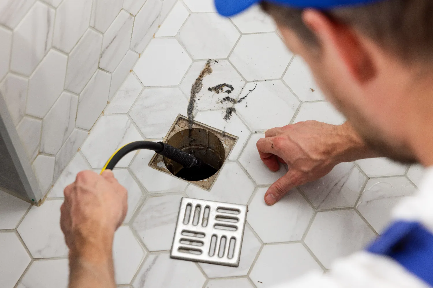 A person demonstrates drain cleaning by using a cable to unclog a floor drain on white hexagonal tile while pointing at the open drain cover.