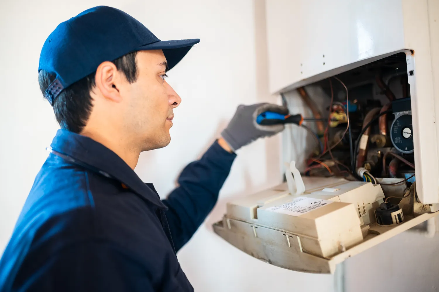 A technician in a blue uniform and cap uses a screwdriver to repair or inspect the wiring inside a wall-mounted water heater, performing expert Water Heater Repair.